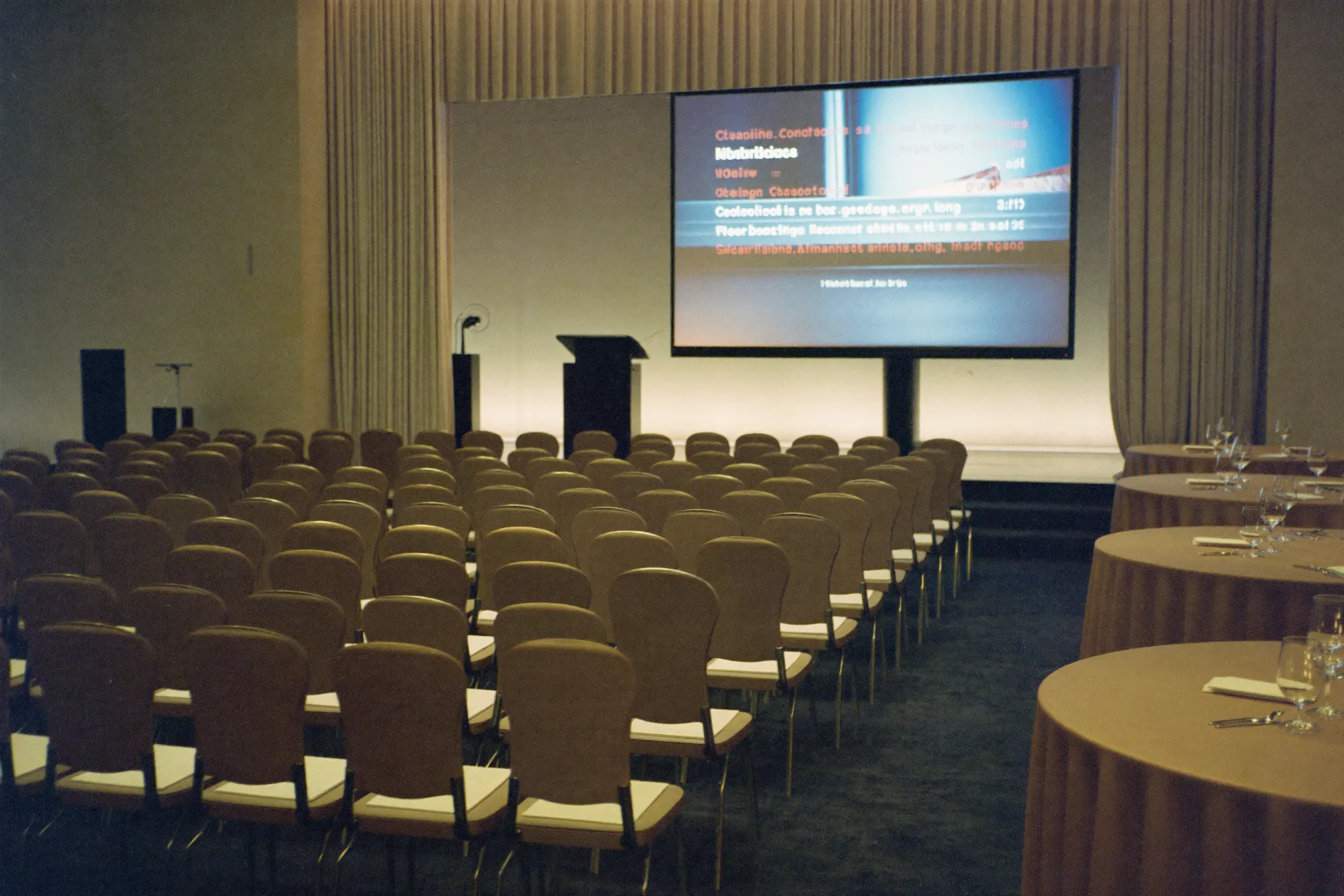 An elegant conference venue set up for a corporate function, with neat rows of chairs facing a stage, a lectern and a large screen, warm ambient uplighting